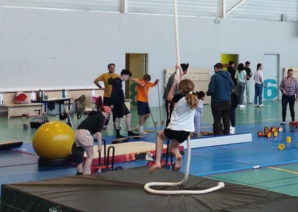 Dans un gymnase, au premier plan, on voit une jeune fille grimper à la corde