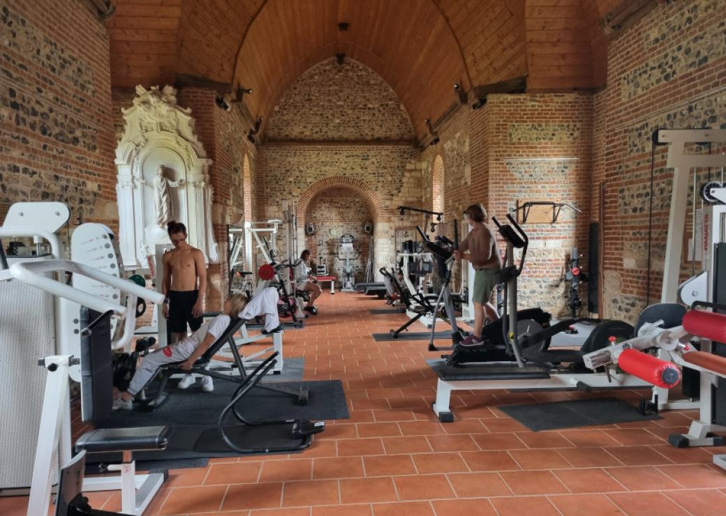 A l'intérieur d'une chappelle, des machines de sport installées de part et d'autres d'une allée centrale, deux personnes debout à côté des machines
