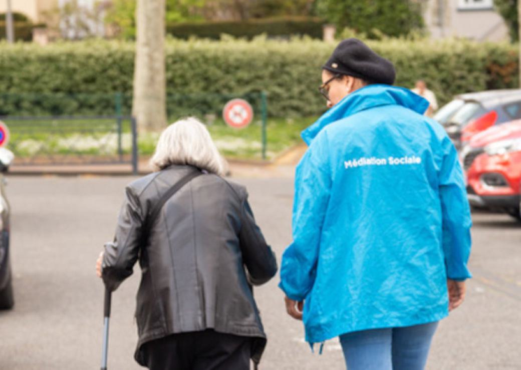 Vue de dos, deux personnes qui marchent dans la rue. L'une d'entre elle porte un blouson bleu sur lequel on peut lire "médiation sociale". L'autre a les cheveux blanc et est équipée d'une béquille