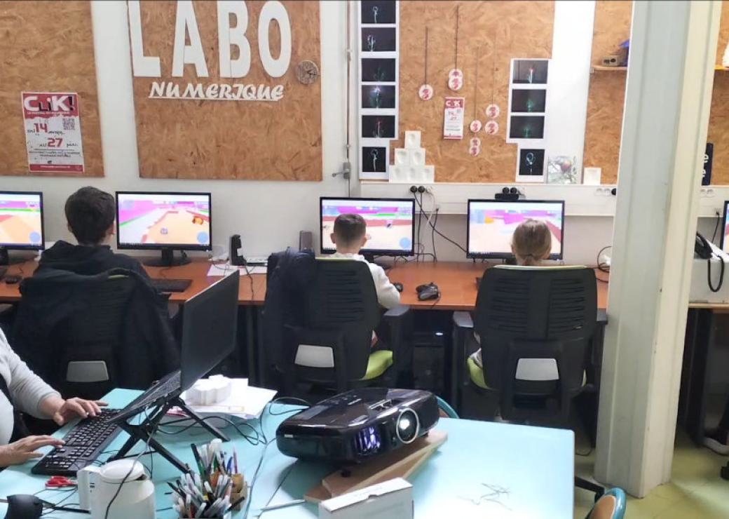 Plusieurs personnes, de dos, attablées face à des écrans. Au mur on peut lire "labo numérique"