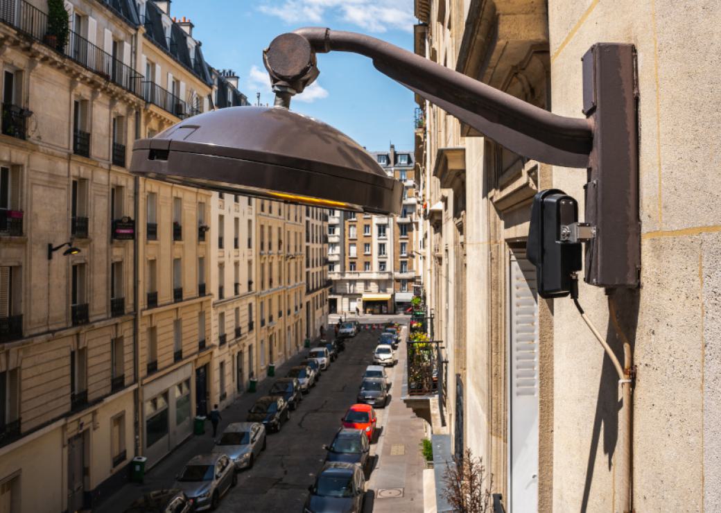 Une tête de lampadaire fixée à un bâtiment  dans rue parisienne. Sous la lampe, un boitier