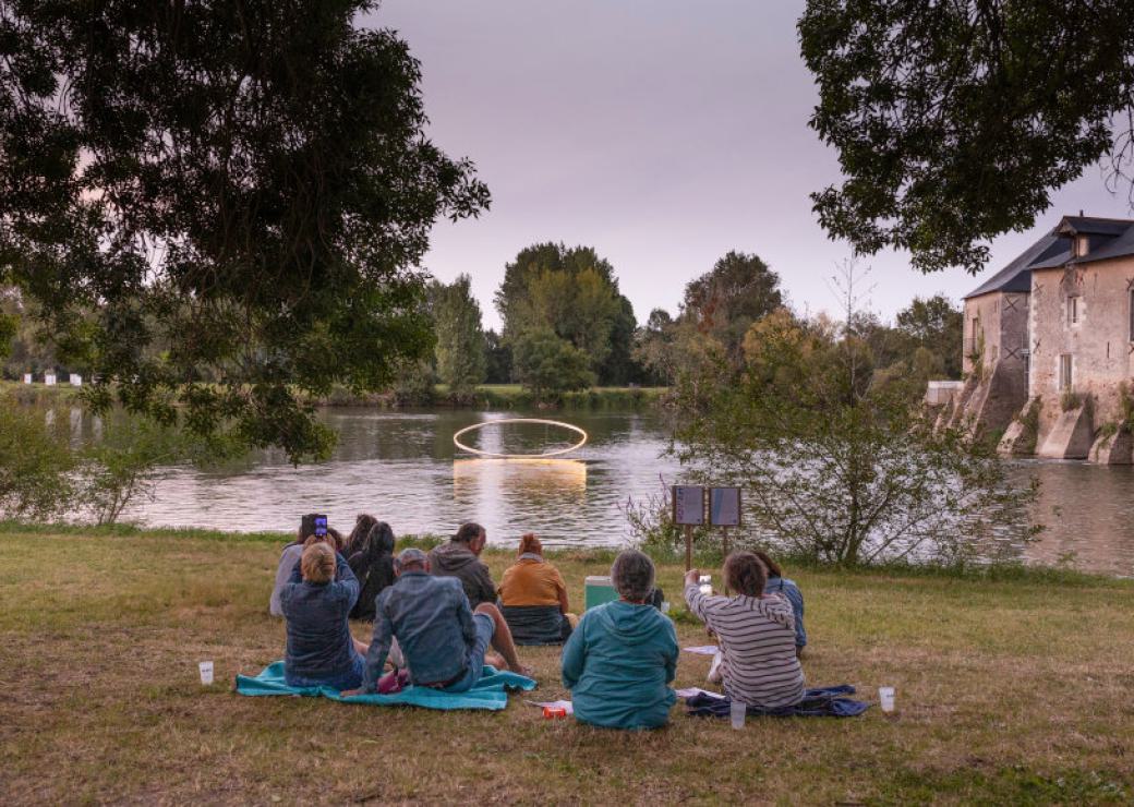 Des personnes assises devant une étendue d'eau sur laquelle flotte une forme sphérique. Au loin, un château.
