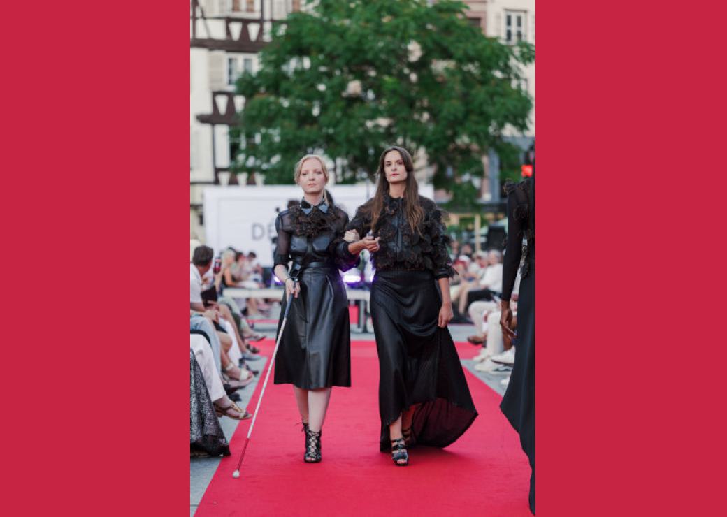 Deux femmes élgantes marchent sur un tapis rouge entouré de public. L'une d'entre elle se dirige avec une canne blanche