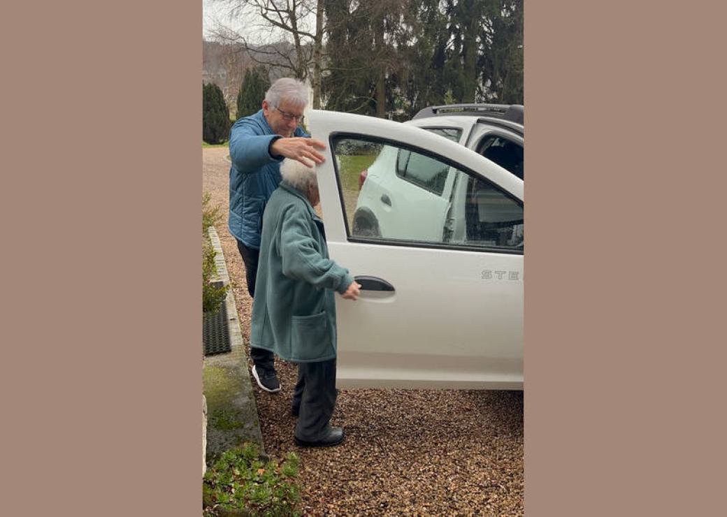 Un monsieur tient une portière de voiture pour laisser s'installer une personne aux cheveux blancs sur le fauteuil passager d'une voiture