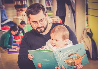 Un petit enfant est installé sur les genoux d'un homme et lit un album dont le titre est attends maman 