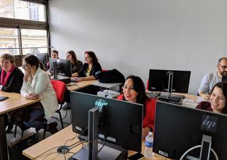 Dans une salle de classe, 8 adultes, principalement des femmes, sont attablés devant des écrans d'ordinateurs