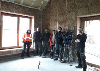 A l'intérieur d'un bâtiment dont les murs sont en paille, 7 personnes posent devant l'objectif du photographe