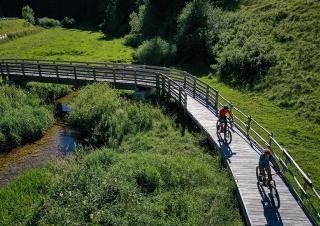 Vue aérienne montrant deux cyclistes roulant sur une passerelle de bois, au dessus d'un paysage très vert, au milieu duquel coule un ruisseau