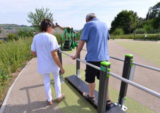 Une dame en blouse blanche accompagne un homme qui marche en se soutenant à deux barres parallèles, sur un terrain équipé d'aménagements de sport