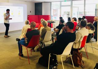 Dans une salle, des personnes assises écoute un homme qui parle au micro, devant une projection de graphiques
