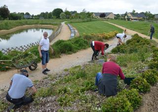 La photo plan large montre 6 personnes en train de travailler la terre d'un espace vert, dans un jardin public où l'on voit aussi un étang sur le côté droit et des maisons en arrière plan
