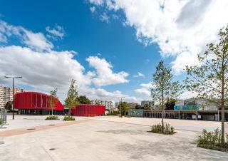 Une large place dallée plantée de quatre jeunes arbres est bordée par un bâtiment moderne rouge et au loin des immeubles d'habitation