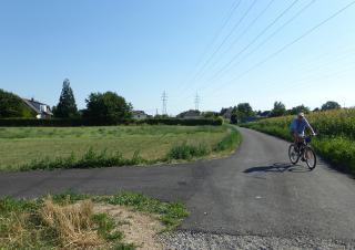 Un homme circule à vélo sur un chemin bordé de champs et de prairie, il avance vers le photographe. Au loin, on distingue une zone pavillonnaire