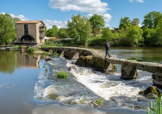 Une personne chemine sur une passerelle de pierre au dessus d'une rivière, en direction d'un moulin sur la berge opposée
