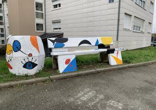 En pied d'immeuble, un banc et un pot de fleur sont décorés avec des motifs colorés, dans le style du peinte miro