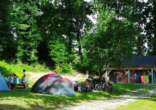 Sous le couvert de grands arbres, deux tentes, des vélos, une petite construction en dur et trois personnes autour d'une table de camping