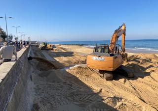 Sur la plage, un tractopelle creuse un chenal, des personnes regardent la scène depuis la promenade