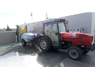 Un tracteur rouge équipé d'une remorque est stationné sur une plateforme de béton. On voit une flaque d'eau au sol.