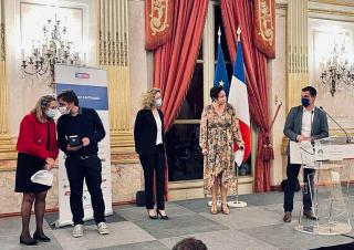 Cinq personnes masquées se tiennent sur une estrade. La scène ressemble à une cérémonie de remise de prix