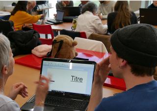 Dans une salle de classe, des personnes font face à des écrans. Au premier blanc, une dame aux cheveux blanc et une jeune personne avec un bonnet regardent le même écran d'ordinateur portable