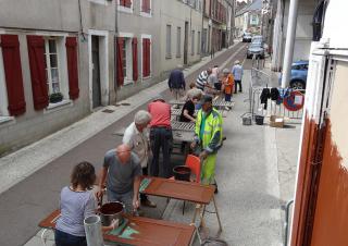 Dans une rue de village, une douzaine de personnes est affairée à peindre des volets de couleur marron