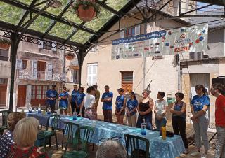 Sur une ^lace de village, des personnes se tiennent en ligne devant une grande table. Ils semblent faire face à un public.