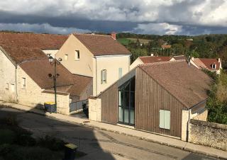 Dans un village, des maisons traditionnelles et un bâtiment rénové avec une grande fenêtre et un bardage de bois sur la façade