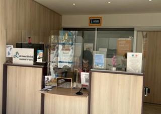 Derrière un comptoir de bois clair surmonté d'une vitre, une dame s'affaire. Au dessus d'elle, un panneau accueil