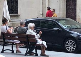 Sur un trottoir, trois personnes sont assises sur un banc face à la rue.