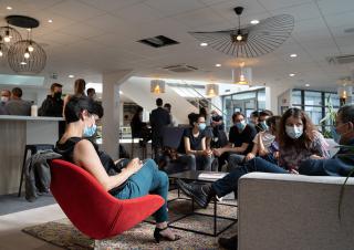 Dans une très grande salle, une quinzaine de personnes portant des masques chirurgicaux sont assises sur des fauteuils bas et des canapés.