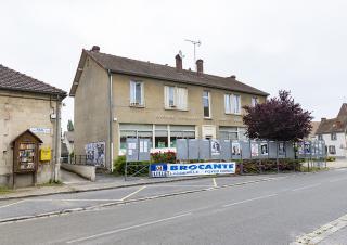 Depuis le bord d'une route, photo d'un bâtiment sur lequel on peut lire groupe scolaire. Une banderole indique aussi Brocante Labbeville Foyer rural 04 juillet  