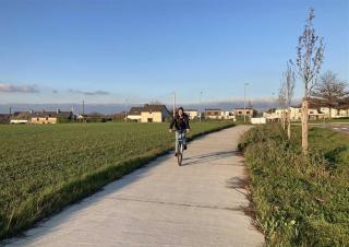 Une personne circule à vélo sur une piste bordée par des champs. Au loin, des pavillons. Ciel bleu