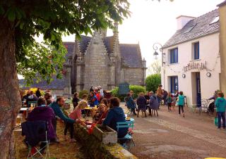 Des gens sont attablés à des tables sur une terrase de café. Un marronnier borde la terrasse, au fond se détache un édifice religieux