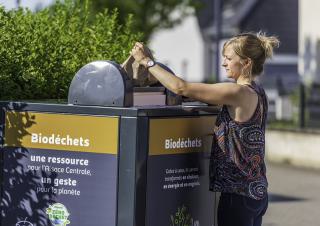 Une femme tient un sac en papier au dessus d'une cuve sur laquelle on peut lire "biodéchets, une ressource pour l'Alsace centrale, un geste pour la planète"