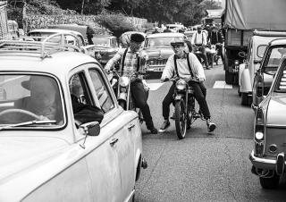 Photo en noir et blanc. Sur une route, un embouteillage de voiture des années cinquante. Au centre, deux motards discutent. Ils portent des bérets et de bretelles