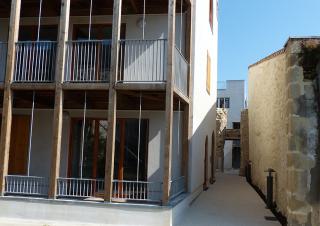 A gauche un batiment de plusieurs étages avec des balcons soutenus par des poutrelles de bois, à droite un mur avec un angle en pierre apparentes, et entre les deux une ruelle étroite