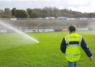 Un homme portant un gilet jaune sur lequel on peut lire "saint-Quentin" marche en direction d'un jet d'eau installé sur une pelouse