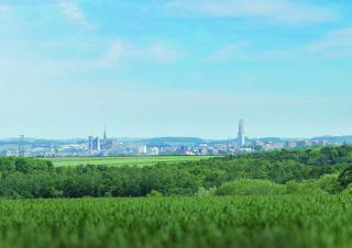 Vue large d'un paysage. Au premier plan, de la végétation, prés et forêts. Au loin, une ville.