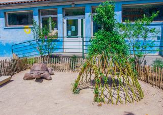 Sur du sable sont posés une tortue en bois et une hutte en roseau. Au fond, un bâtiment bleu