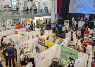 Vue d'en haut, une vaste salle cloisonnée en petits stands. Des personnes évoluent dans ces stands