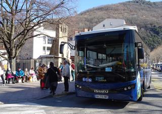 Dans une ville, un autocar est arrêté devant un abribus où sont assises des personnes. En arrière plan, une colline boisée