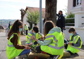 Des enfants habillés de gilets jaunes plantent des végétaux au pied d'un arbre sur un trottoir