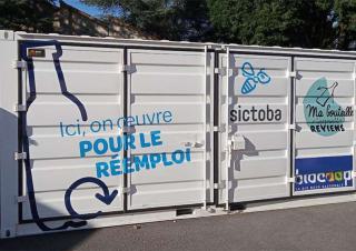 Un container blanc sur lequel on peut lire "ici on œuvre pour le réemploi", "sictoba" et "ma bouteille s'appelle reviens"