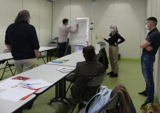 5 personnes dans une salle de réunion. Certaines portent un masque chirurgical. L'une d'entre elle écrit sur un tableau blanc.