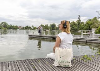 Une femme vetue de blanc est assise sur un ponton de bois,, face à une étendue d'eau. Elle porte un casque audio sur les oreilles