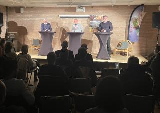 Dans une salle, trois personnes se tiennent sur une estrade devant un public, face à des personnes assises