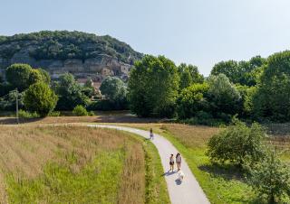 Vu d'en haut, un chemin sur lequel se promènent deux personnes et leur chien et une personne à vélo. Paysage de campagne