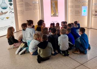 Une groupe d'une quinzaine d'enfants est assis sur le sol, un adulte semble leur parler. Ils sont entourés de panneaux lumineux avec des œuvres d'art