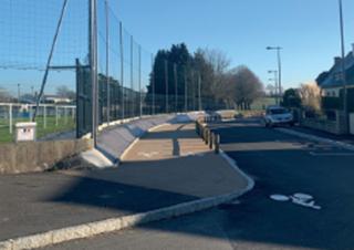 Une route longée par un lotissement et par un terrain de sport. Elle est bordée par une piste cyclable