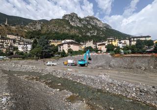 Des engins de travaux sur les berges d'une rivière. En fond, un village bordé par une montagne